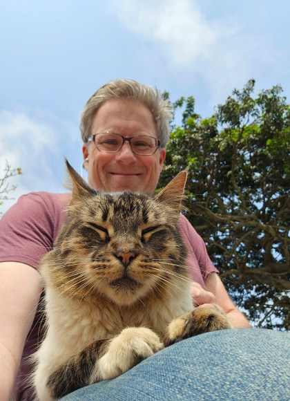 Photo of author Jeff Bogle, sitting with his cat in his lap by a tree. Jeff and the cat are facing the camera, angled down. Jeff is smiling. The cat’s eyes are closed; it looks content.