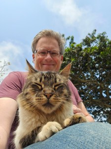 Photo of author Jeff Bogle, sitting with his cat in his lap by a tree. Jeff and the cat are facing the camera, angled down. Jeff is smiling. The cat’s eyes are closed; it looks content.